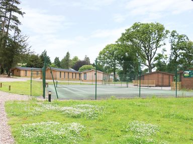 An outdoor tennis court surrounded by a green fence with wooden buildings and trees in the background at Lodge 8 Louth