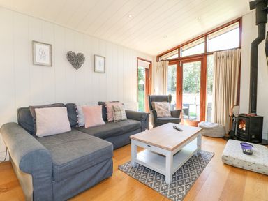 A living room with a gray sofa, armchair, wooden coffee table, rug, wood stove, and large windows with curtains at Brooke in Exton Rutland