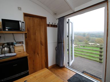 An open door leading to a wooden deck with a view of sheep in a fenced grassy area from inside a kitchen with microwave kettle and wooden cabinets at Shepherds Cabin at Titterstone in Clee Hill
