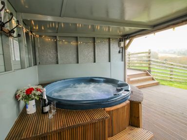 An outdoor covered patio with a hot tub flowers champagne and glasses on a wooden deck at Shepherds Cabin at Titterstone in Clee Hill
