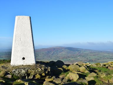 A white trig point on rocky ground with a view of fields and hills in the background at Shepherds Cabin at Titterstone in Clee Hill