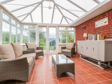 A conservatory with wicker chairs and a sofa around a glass table and a cabinet with books at Bower View in Madley
