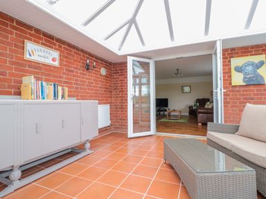 A sunroom with terracotta floor tiles a gray cabinet with books a wicker sofa and glass coffee table leading to a living room at Bower View in Madley