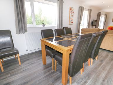 A dining area with a wooden table and six black leather chairs near windows with grey curtains at Heron Lodge in Mavis Enderby near Spilsby