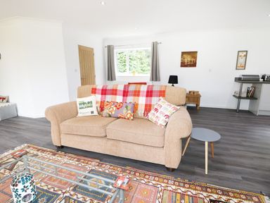 A living room with a beige sofa with cushions and a plaid throw a small round side table a patterned rug and a glass coffee table at Heron Lodge in Mavis Enderby near Spilsby