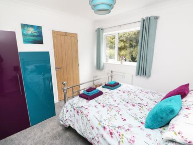 A bedroom with a floral bedspread, pillows, folded towels, a wooden door, wardrobe with purple and blue doors, window with curtains and a painting on the wall at Heron Lodge in Mavis Enderby near Spilsby