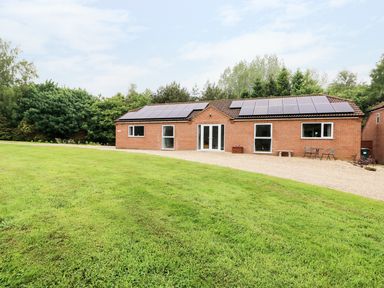 A single story brick building with solar panels on the roof facing a gravel driveway and lawn surrounded by trees at Heron Lodge in Mavis Enderby near Spilsby