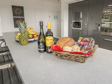 A kitchen island with a basket of bread and snacks a bottle of wine a bottle of prosecco and a fruit bowl with pineapple bananas and other fruits at Manor House in Warkworth