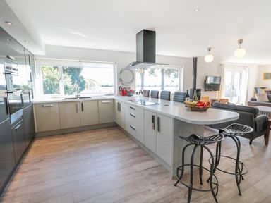 A kitchen with an island, bar stools, built-in ovens, large windows, and an open living area at Manor House in Warkworth