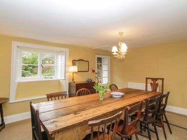 A dining room with a large wooden table surrounded by chairs tulips in a vase on the table and a window with a view at Crannacombe Farmhouse in Loddiswell