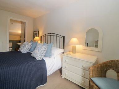 A bedroom with a bed with blue pillows and blanket a white dresser with a lamp and mirror a wicker chair and an open door leading to a bathroom at Crannacombe Farmhouse in Loddiswell