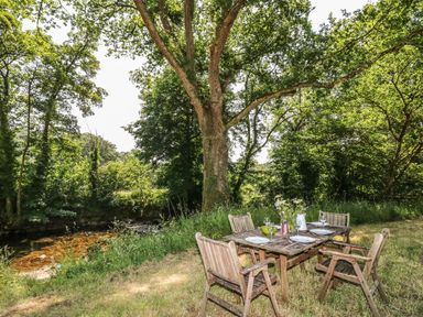 An outdoor wooden table with four chairs set with plates and glasses near a tree and a small stream at Crannacombe Farmhouse in Loddiswell