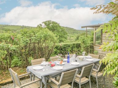 An outdoor dining table with chairs set with plates glasses and bottles near bushes and hills at Crannacombe Farmhouse in Loddiswell