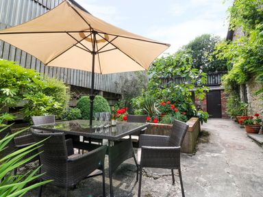 An outdoor patio with a table and chairs under a large umbrella surrounded by plants and flowers at The Cider Barn in Loddiswell