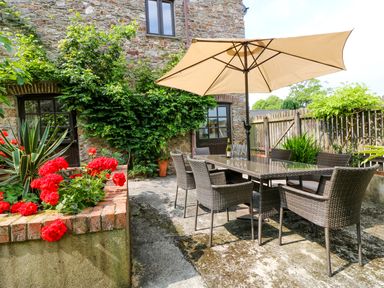 An outdoor patio with a glass dining table and six wicker chairs under a beige umbrella next to a stone house with plants and red flowers at The Cider Barn in Loddiswell