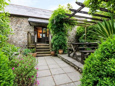 A stone patio with wooden steps leading to a door and a pergola with a picnic table surrounded by plants at The Cider Barn in Loddiswell