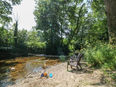 A riverbank with two chairs and children's sand toys near a flowing river surrounded by trees at The Cider Barn in Loddiswell