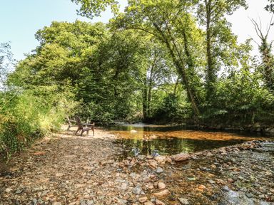 A rocky riverbank with two chairs and glasses near a flowing river surrounded by trees at The Cider Barn in Loddiswell