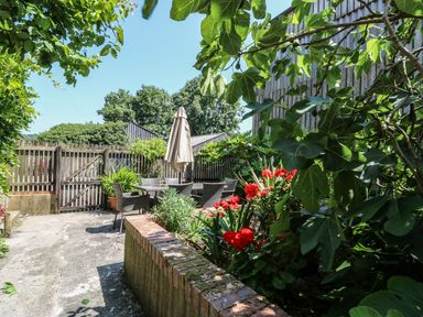 An outdoor patio area with a wooden table and chairs under a closed umbrella surrounded by plants and flowers at The Cider Barn in Loddiswell