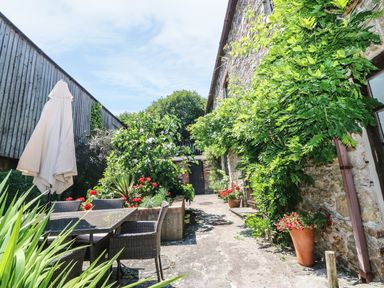 An outdoor patio area with a glass table and wicker chairs surrounded by plants and flowers at The Cider Barn in Loddiswell