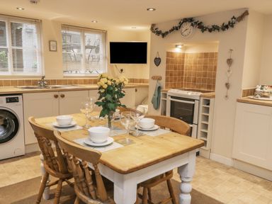 A kitchen with a wooden dining table and chairs set with bowls and glasses a washing machine under the counter a stove a wall clock and windows with blinds at Robins Nest in Whitby