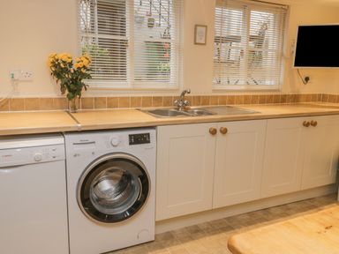 A kitchen counter with a sink washing machine dishwasher flowers in a vase under windows with blinds and a TV mounted on the wall at Robins Nest in Whitby