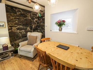 A dining area with a wooden table and chairs a beige armchair next to a stone wall and a window with flowers on the sill at Ty Coed in Llanberis