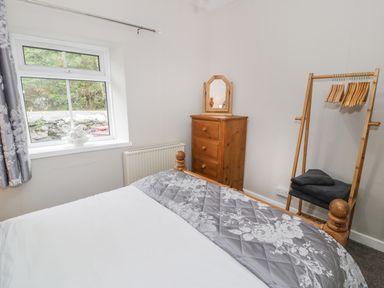 A bedroom with a bed with a grey floral blanket a wooden chest of drawers with a mirror and a wooden clothes rack at Ty Coed in Llanberis
