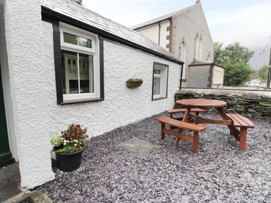 An outdoor area with a white building, two windows, a wooden picnic table, a flower pot, and a stone wall at Ty Coed in Llanberis
