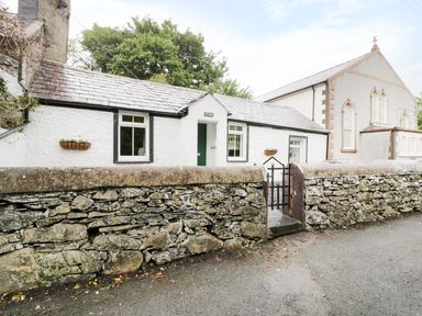 A white stone cottage with a green door behind a low stone wall and metal gate at Ty Coed in Llanberis