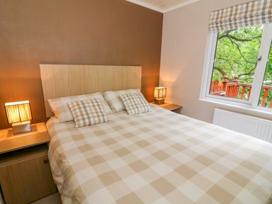 A bedroom with a double bed with checkered bedding and pillows two bedside tables with lamps and a window with a view of trees at Willow Lodge in Troutbeck Bridge