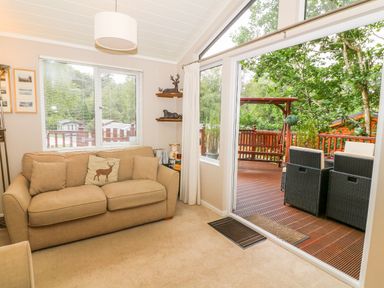 A living room with a sofa and deer cushion next to a window and glass door leading to a wooden deck with outdoor furniture at Willow Lodge in Troutbeck Bridge