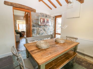 A dining area with a wooden table, two woven placemats, chairs, a bench, stone wall with a cow painting, and doorway to a living room at Beudy in Chwilog