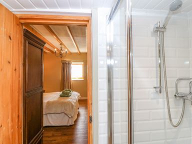 A bathroom with a shower area showing a handheld showerhead and white tiled walls next to a doorway leading to a bedroom with a bed with towels on it and a window with curtains at Beudy in Chwilog