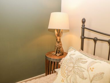 A bedside table with a lamp featuring a rabbit base next to a bed with a metal headboard and a pillow with a floral design at Beudy in Chwilog