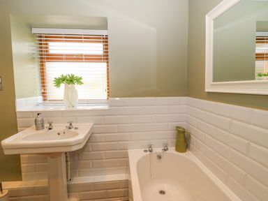 A bathroom with a white sink and bathtub a green jug on the tub edge and a window with blinds at Beudy in Chwilog
