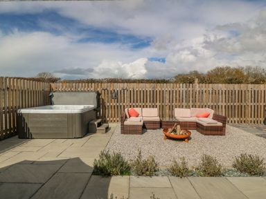 An outdoor patio area with a hot tub wicker seating with cushions and a fire pit at Beudy in Chwilog