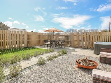 An outdoor patio with a table and chairs under an umbrella a fire pit and a hot tub in a fenced yard at Beudy in Chwilog