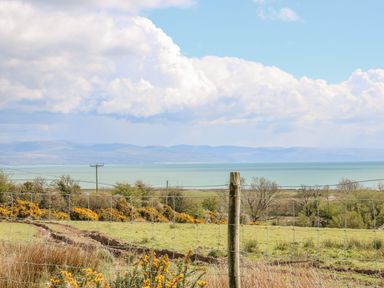 A fenced grassy field with yellow flowers and trees overlooking a large body of water and distant mountains at Beudy in Chwilog