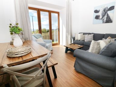 A living room with a wooden dining table with chairs a vase with flowers a blue armchair a blue sofa with cushions a small wooden coffee table and a painting of a cow on the wall at Sgubor in Chwilog