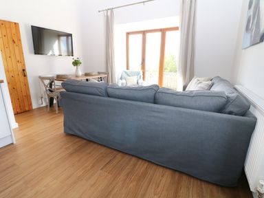 A living room with a gray sofa a wooden dining table with chairs a wall mounted television and glass doors at Sgubor in Chwilog