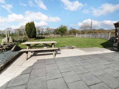 A backyard with a wooden picnic table on a paved patio and a lawn enclosed by a wooden fence and stone walls at Sgubor in Chwilog