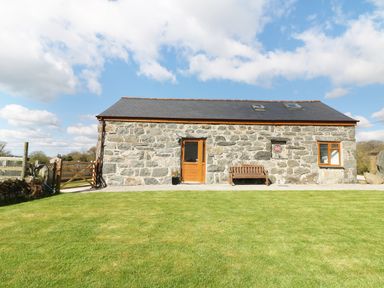 A stone cottage with a wooden door and bench on a lawn under a partly cloudy sky at Sgubor in Chwilog