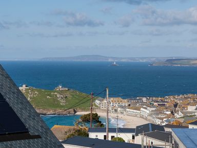 A coastal town with rooftops and a beach overlooking the ocean with a lighthouse on a small island at Godrevy in St Ives