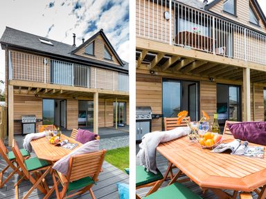 An outdoor wooden dining table with chairs cushions flowers fruit and a magazine on a deck outside a wooden house with balcony and barbecue at Godrevy in St Ives