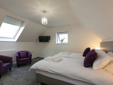 A bedroom with two single beds white bedding purple pillows two purple armchairs a wall mounted TV and skylight window at Godrevy in St Ives