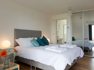 A bedroom with a bed with white linens and teal pillows a bedside table with a lamp and flowers and a mirrored closet at Godrevy in St Ives