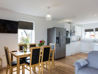 A dining area with wooden table and chairs next to a modern kitchen with white cabinets and stainless steel appliances at Godrevy in St Ives