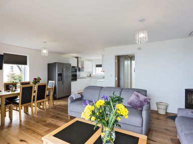 An open plan living area with a wooden dining table and chairs a kitchen with white cabinets and a refrigerator and a seating area with a gray sofa and flowers on a coffee table at Godrevy in St Ives