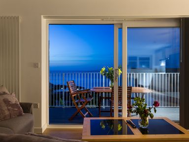 A living room with a sofa flowers on a table and a balcony with wooden chairs and a table overlooking the sea at Godrevy in St Ives